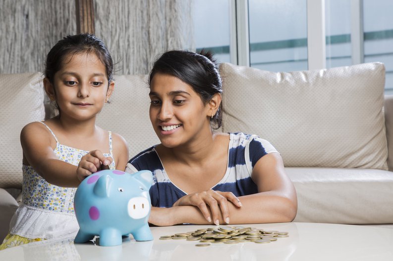 A young girl putting coins in her piggy bank as her mom looks on proudly.