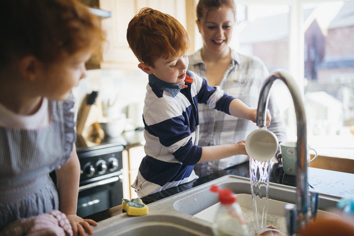 A mother and her two young children are washing dishes in a bright kitchen.