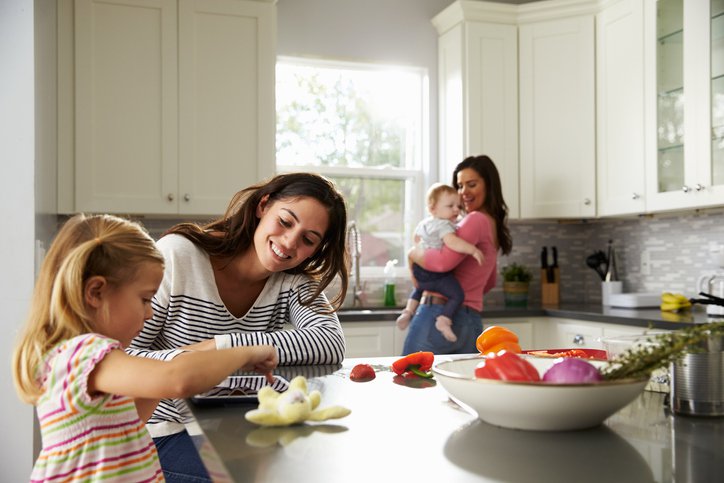 A mom playing with her daughter at a kitchen counter while the other mom dances with their baby in the background.