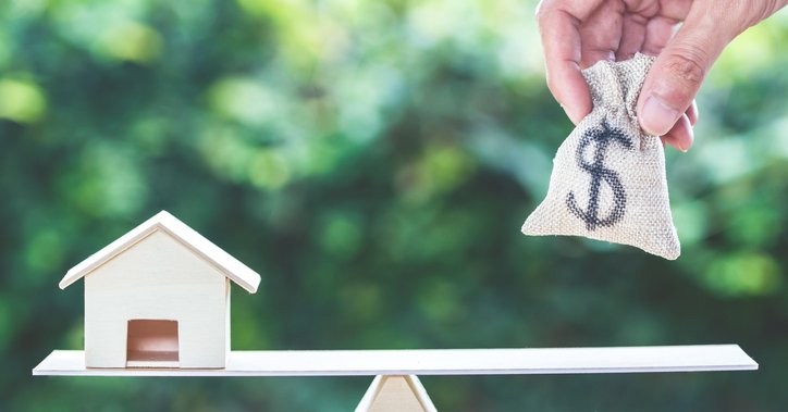 A hand places a bag with a dollar sign onto scales with a toy house on the other side.