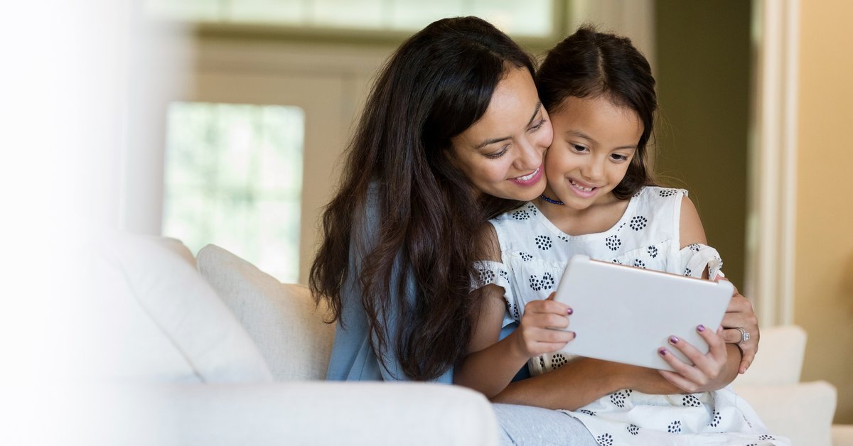 A mother and her young daughter cuddling on the couch while looking at a tablet computer.