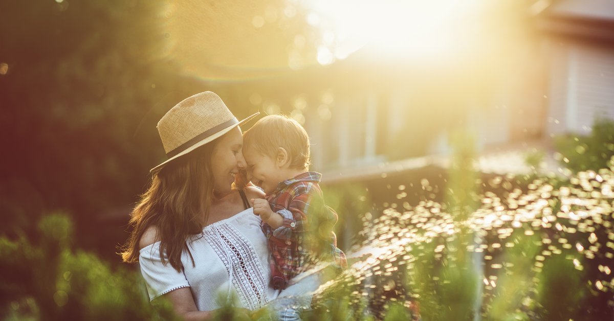 A mother holding her young son while watering plants in the backyard.