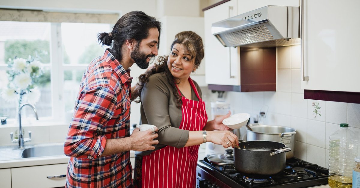 A mother cooking at the stove with her adult son looking over her shoulder.