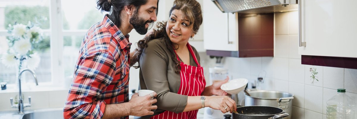 A mother cooking at the stove with her adult son looking over her shoulder.