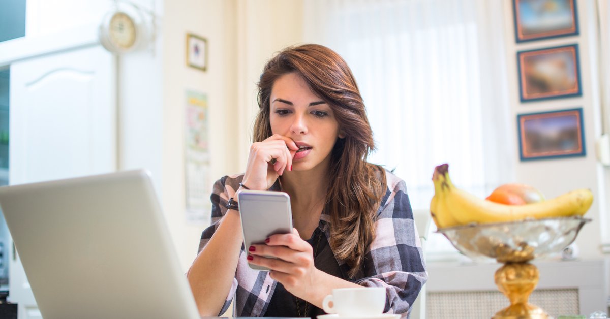 A nervous young woman biting her fingernail while sitting in her kitchen with her phone and laptop.