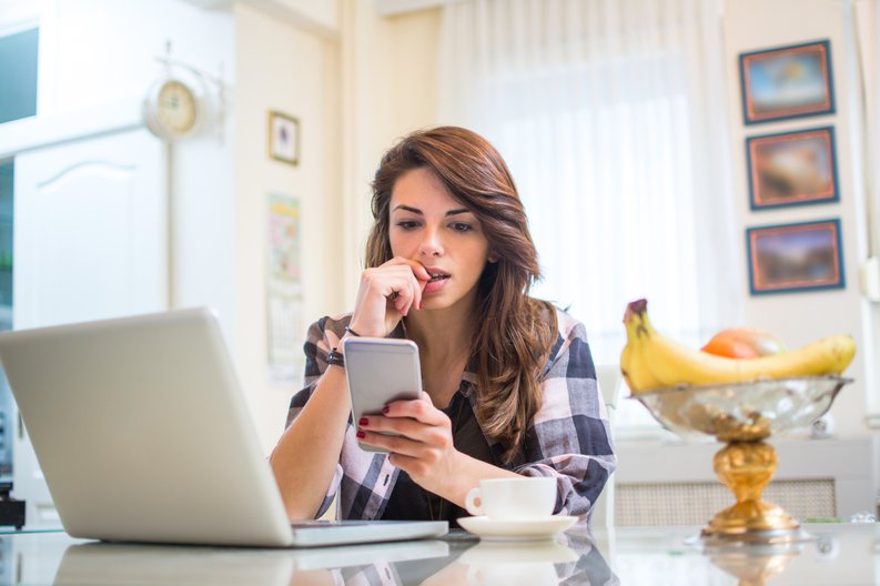A nervous young woman biting her fingernail while sitting in her kitchen with her phone and laptop.
