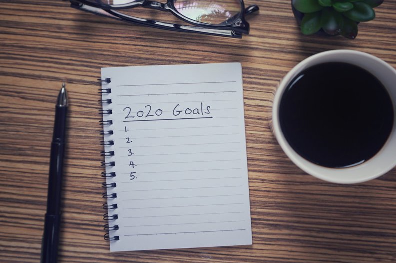 A wooden desk with a pen, cup of coffee, and a notebook that has the words 2020 Goals and a numbered list written on a page.