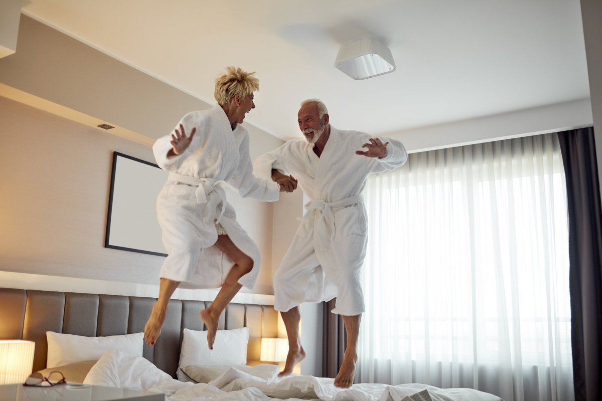 An older couple jumping on the bed in hotel bathrobes.