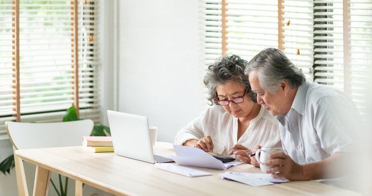 An older man and woman sitting at their dining table and looking through paper bills with a laptop open in front of them.