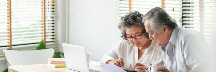 An older man and woman sitting at their dining table and looking through paper bills with a laptop open in front of them.