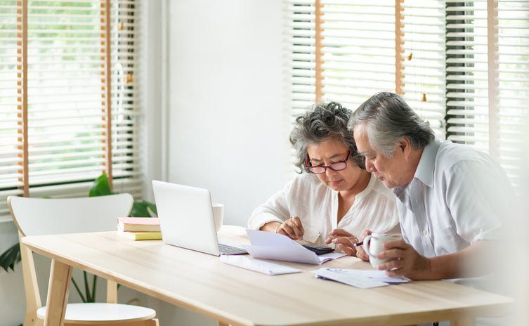 An older man and woman sitting at their dining table looking at paper bills with an open laptop in front of them.