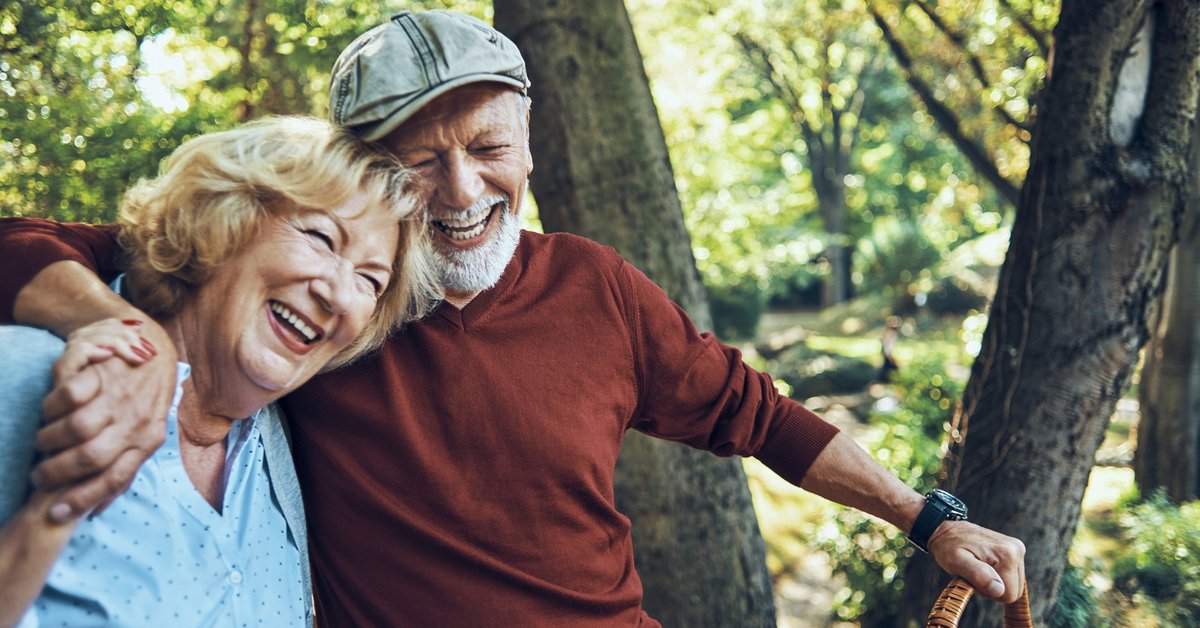 An older couple smiling with their arms around each other and holding a picnic basket in the woods.