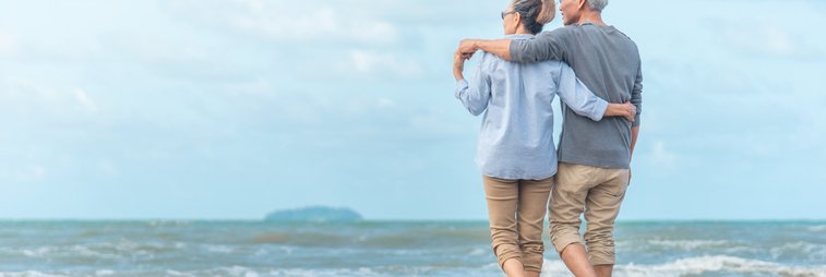 An older couple walking along the beach with their arms around each other.