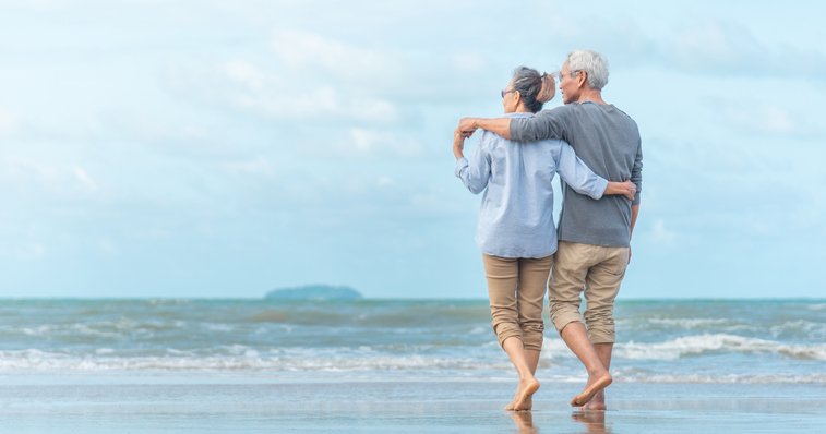 An older couple walking along the beach with their arms around each other.