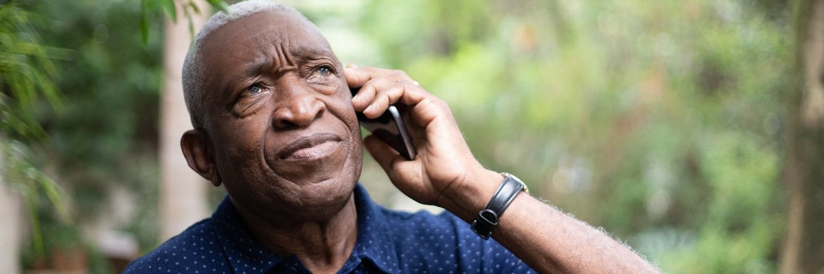 An older man standing under green trees while making a phone call.
