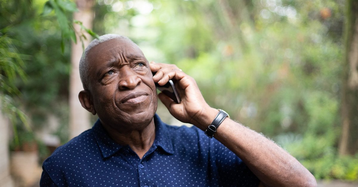 An older man standing under green trees while making a phone call.