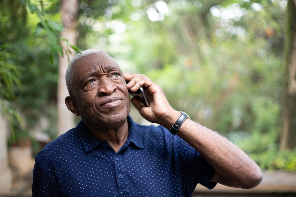An older man standing under green trees while making a phone call.