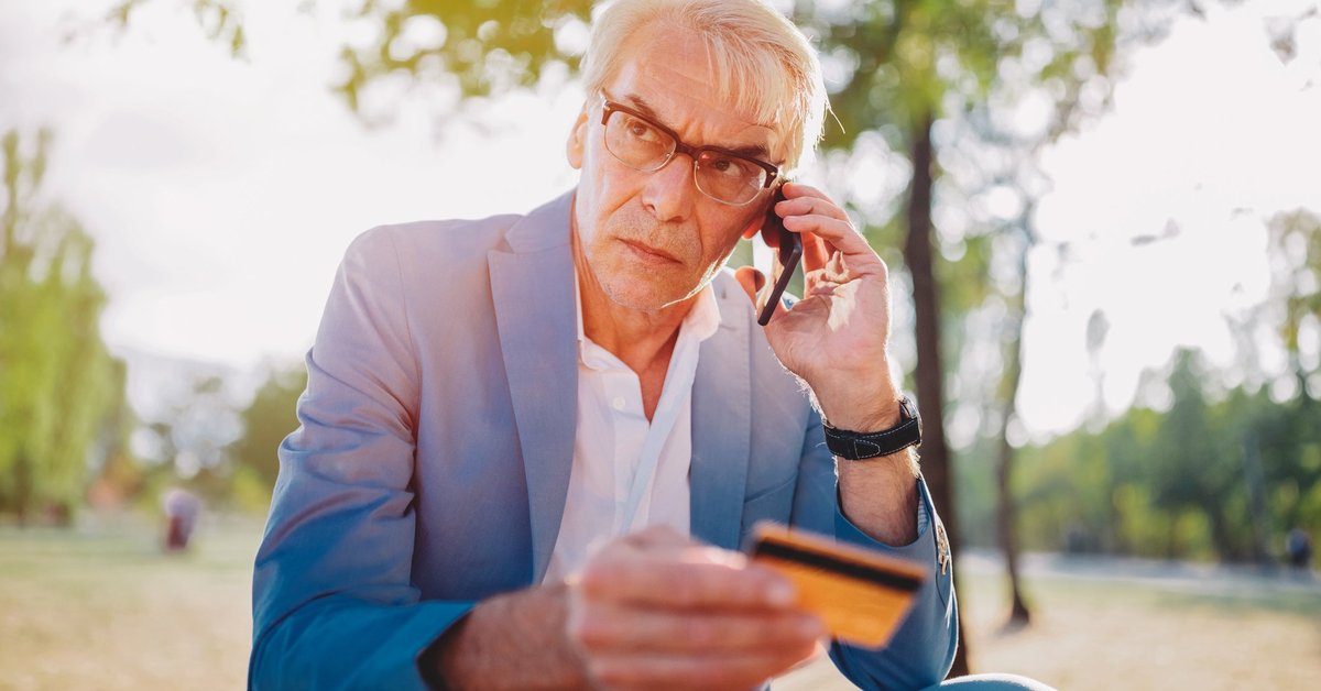 A distraught man sitting in a park and talking on his phone while holding a credit card.