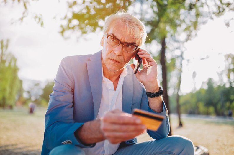 A distraught man sitting in a park and talking on his phone while holding a credit card.