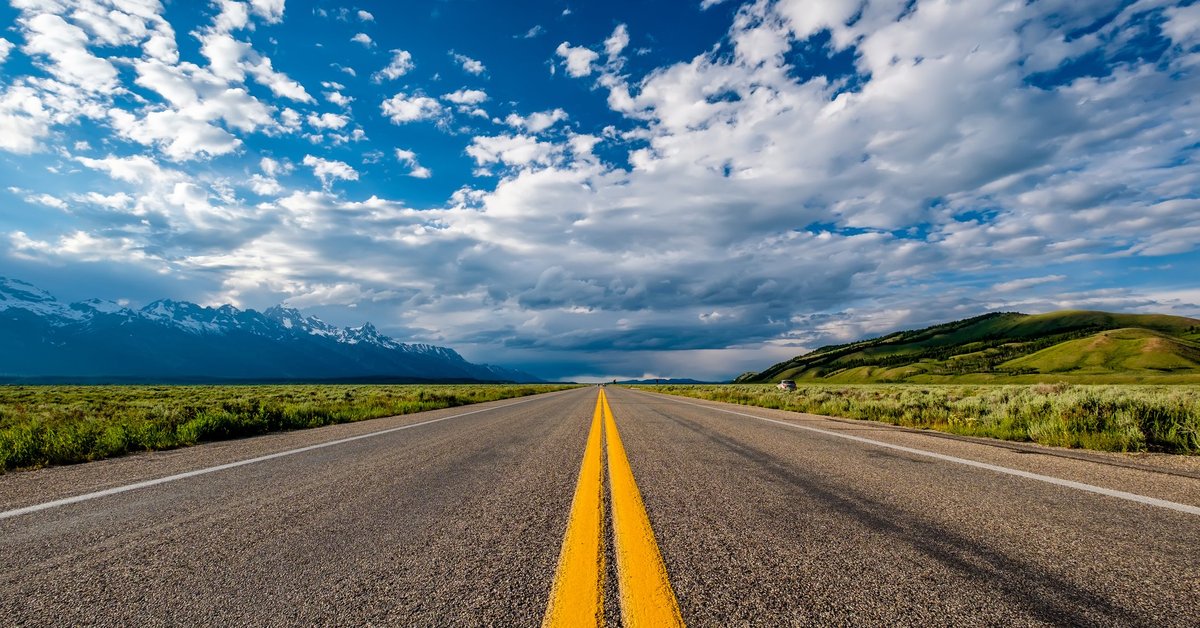 open road in Wyoming with green hills on one side and snow-capped mountains on the other