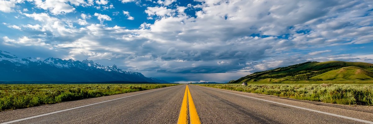 open road in Wyoming with green hills on one side and snow-capped mountains on the other