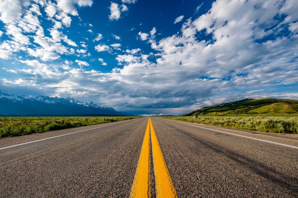 open road in Wyoming with green hills on one side and snow-capped mountains on the other