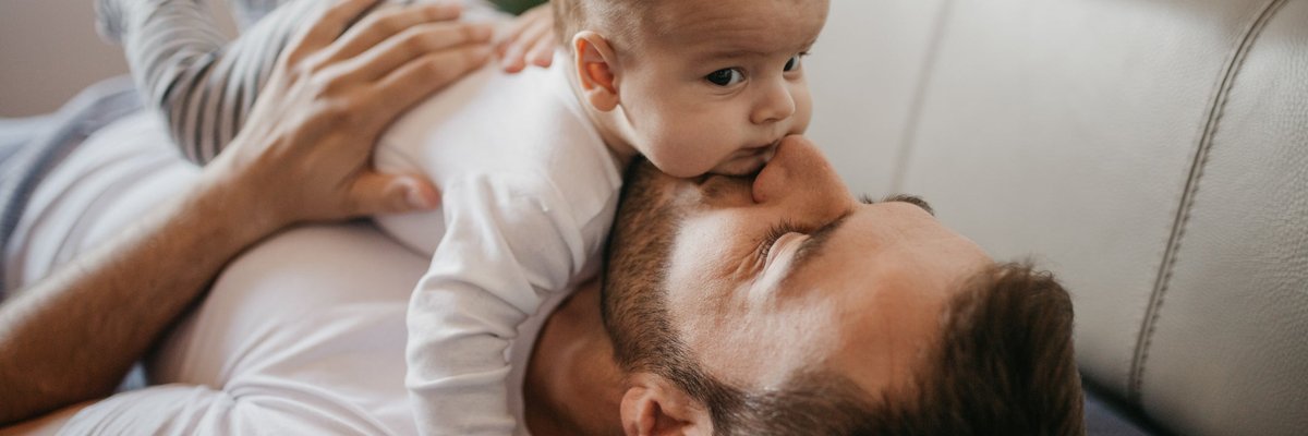A parent lying on the couch and kissing their young baby lying on top of them.