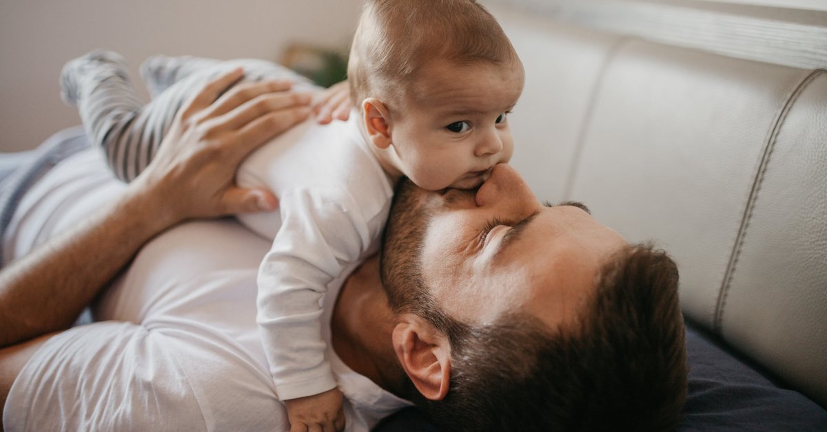 A parent lying on the couch and kissing their young baby lying on top of them.