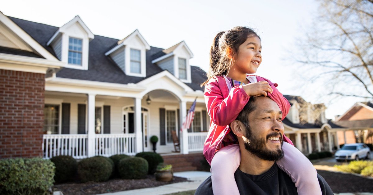 A parent standing in the front yard of their house with their young child sitting on their shoulders.