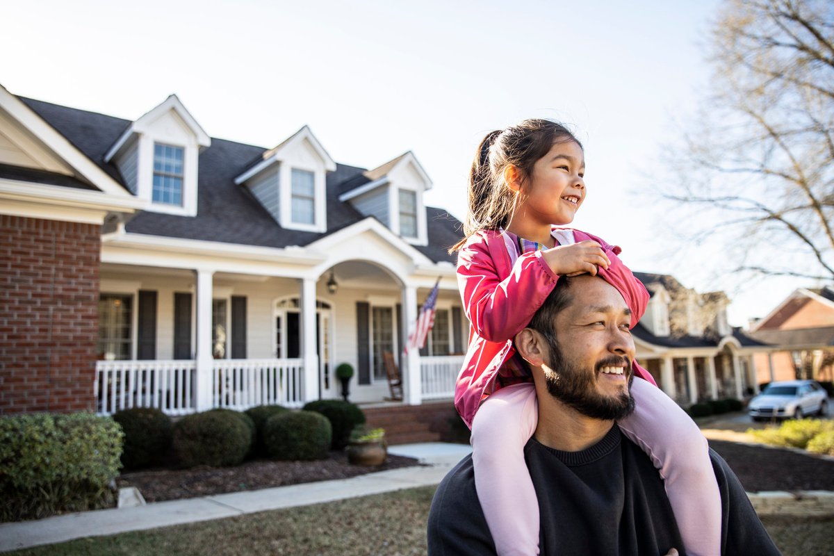 A parent standing in the front yard of their house with their young child sitting on their shoulders.