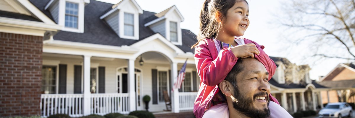 A parent standing in the front yard of their house with their young child sitting on their shoulders.
