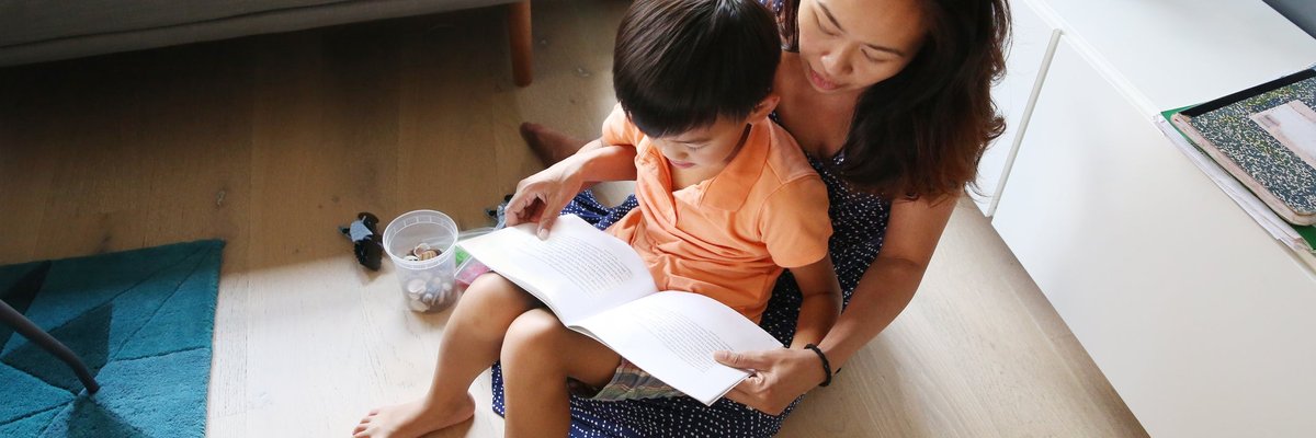 A parent reading a book to their young child sitting in their lap on their living room floor.