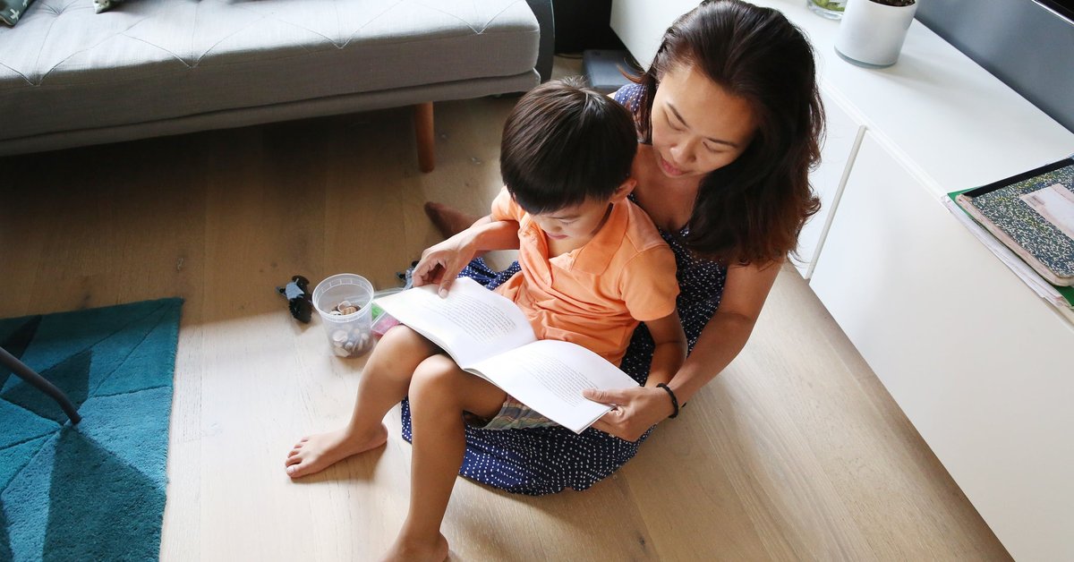 A parent reading a book to their young child sitting in their lap on their living room floor.