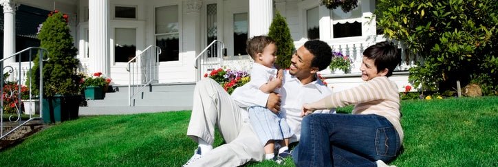 A mom and dad with their baby sitting on the sunny lawn in front of their house.