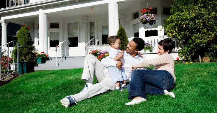 A mom and dad with their baby sitting on the sunny lawn in front of their house.