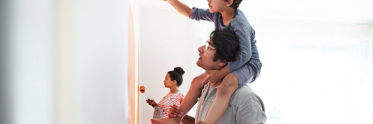 A child sitting on his parent's shoulders and painting the walls of a room while the other parent paints behind them.
