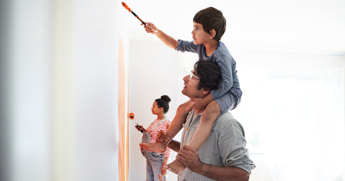 A child sitting on his parent's shoulders and painting the walls of a room while the other parent paints behind them.