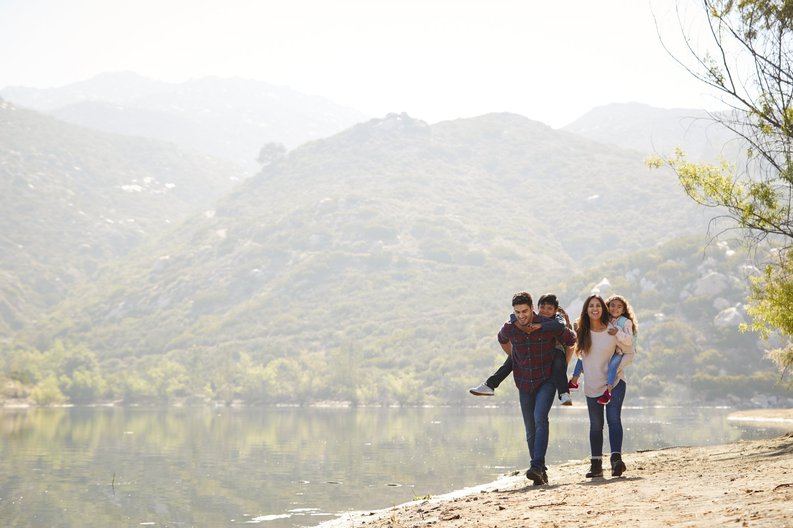 Two young kids piggybacking on their parents as they hike next to a lake with hills in the background.