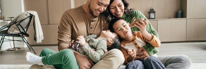 A smiling mom and dad sitting on the floor with their young kids playing in their laps.