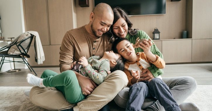 A smiling mom and dad sitting on the floor with their young kids playing in their laps.