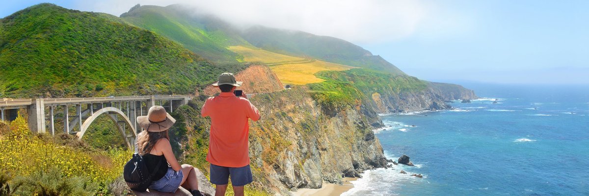 Two people standing near the edge of an ocean cliff and admiring the view of a bridge and the surrounding nature area.