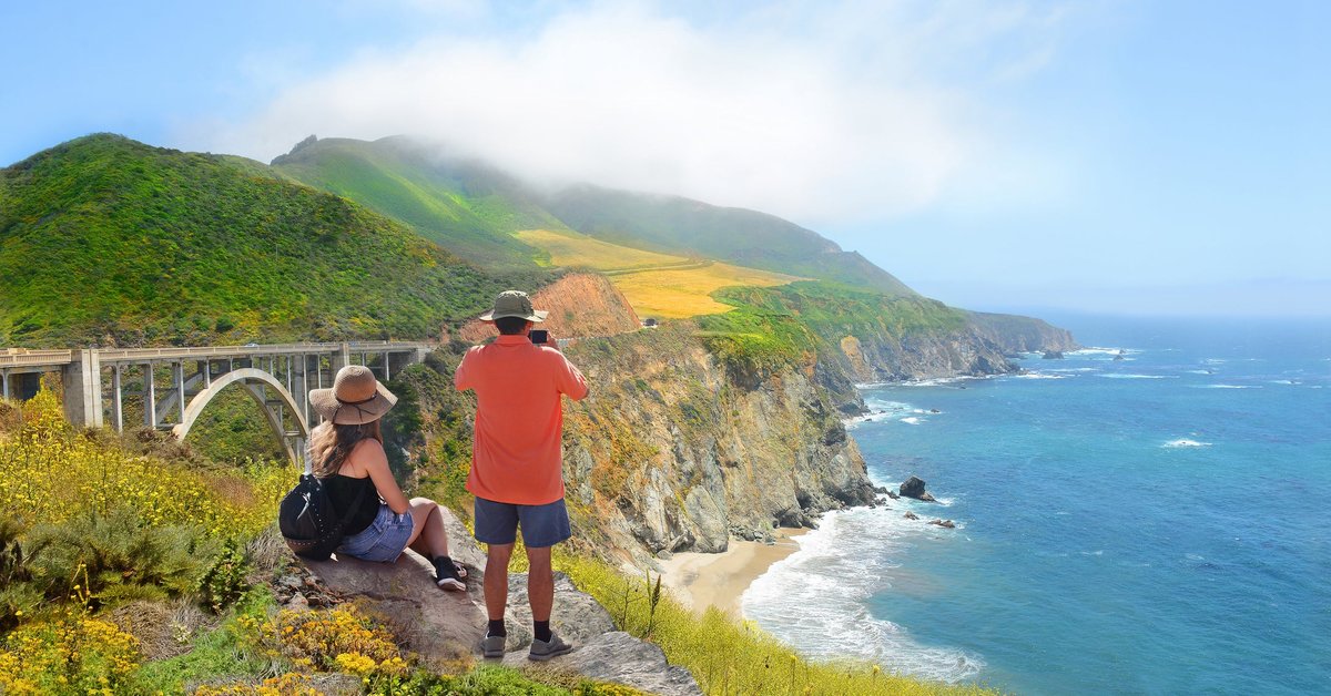 Two people standing near the edge of an ocean cliff and admiring the view of a bridge and the surrounding nature area.
