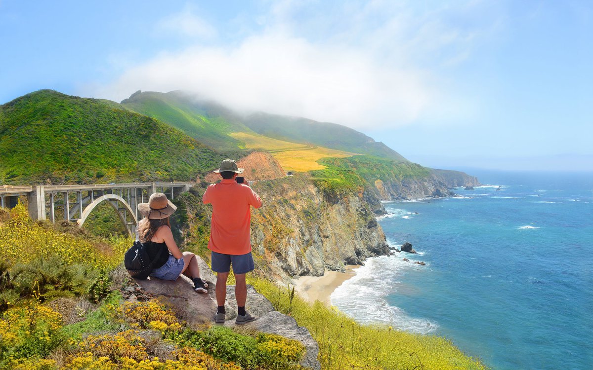 Two people standing near the edge of an ocean cliff and admiring the view of a bridge and the surrounding nature area.