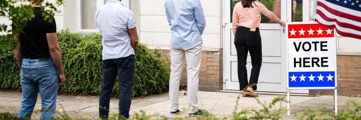 People standing in line outside a building next to a sign that says Vote Here below an American flag.