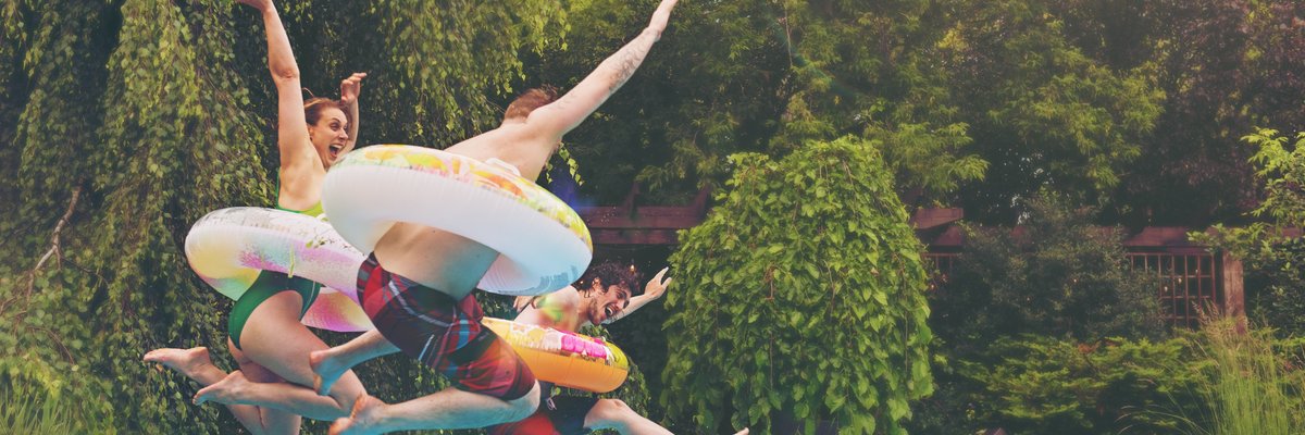 Three people wearing colorful tubes around their waists jumping into a backyard pool surrounded by trees.