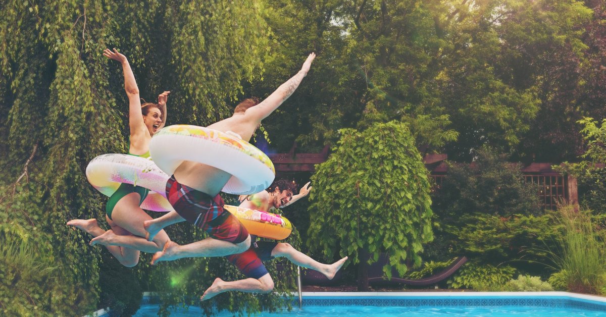 Three people wearing colorful tubes around their waists jumping into a backyard pool surrounded by trees.