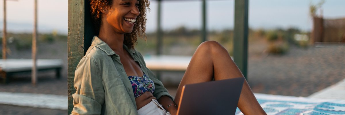 A smiling person sitting on a cabana bed on the beach while typing on a laptop.