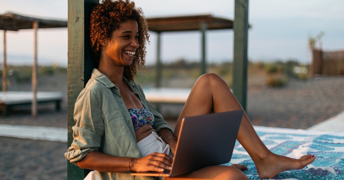 A smiling person sitting on a cabana bed on the beach while typing on a laptop.