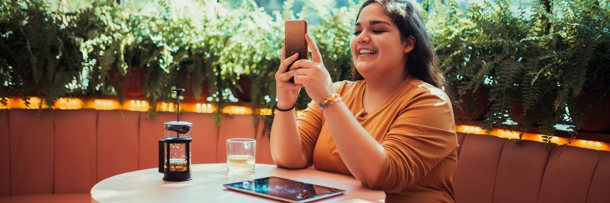 A smiling person sitting at a table in a coffee shop and looking at a phone in their hand