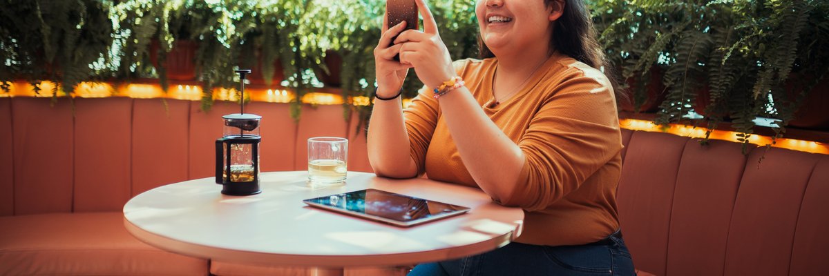 A smiling person sitting at a table in a coffee shop and looking at a phone in their hand